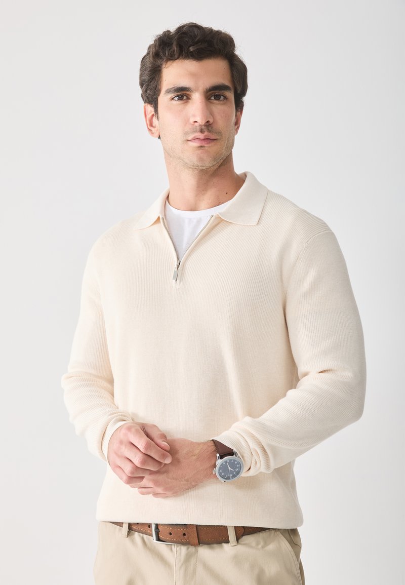 Sudadera de media cremallera color beige claro, con textura acanalada, mangas largas, cuello con solapa, usada sobre una camiseta blanca, combinada con pantalones beige y un cinturón marrón.