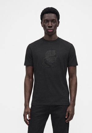 Mand iført en sort t-shirt med korte ærmer og et diskret tekstureret silhuetgrafik på brystet, stående mod en ensfarvet hvid baggrund.