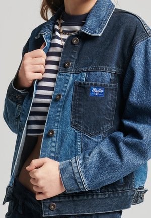 Osoba nosząca dwukolorową dżinsową kurtkę na paskowanej koszuli, trzymająca kołnierz kurtki jedną ręką i regulująca dół kurtki drugą.