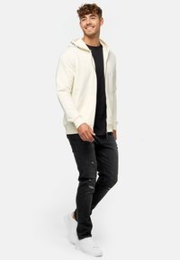 Crème zip-up hoodie met een zwarte accent op de kraag, gecombineerd met versleten zwarte jeans en witte sneakers. Eenvoudig, casual ontwerp.