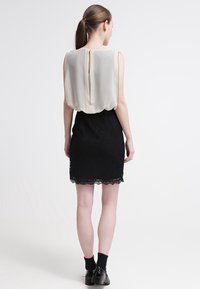 Femme aux cheveux bruns en queue de cheval, portant un blouse sans manches blanche rentrée dans une jupe noire à dentelle et des bottines noires.