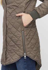 Abrigo acolchado e insulated en verde olivo con un patrón diagonal. Cuenta con un bolsillo con cremallera lateral y puños acanalados; el dobladillo es ligeramente más largo en la parte posterior.