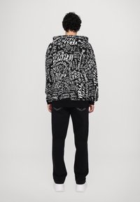 Hoodie noir avec un design de texte blanc, doté d'une poche avant et d'une capuche à cordon. Accompagné de pantalons noirs et de baskets blanches.