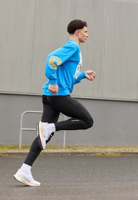 Blue sweatshirt with printed design, black leggings, and white athletic shoes with orange accents, worn by a male runner in motion.