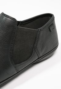 Chaussure à enfiler en cuir noir avec panneau élastique sur le côté et surface texturée, présentée en gros plan incliné.