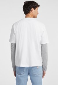 Camiseta blanca de algodón de manga corta, superpuesta sobre una camisa de manga larga gris. Jeans de mezclilla azul claro con un pequeño logotipo de la marca en la parte posterior.