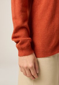 Pull en laine couleur rouille avec des poignets côtelés, associé à un tissu beige clair. Présente une texture douce et une coupe décontractée.