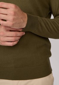 Pull en maille vert olive avec des poignets et un ourlet côtelés. Gros plan sur des mains tenant le pull, mettant en avant la texture et la coupe.