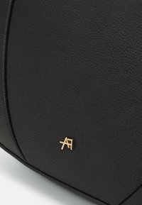 Czarna torba z teksturowanej sztucznej skóry z akcentem w postaci złotego metalowego logo. Charakteryzuje się ustrukturyzowanym designem i widocznymi szwami dla podkreślenia detali.
