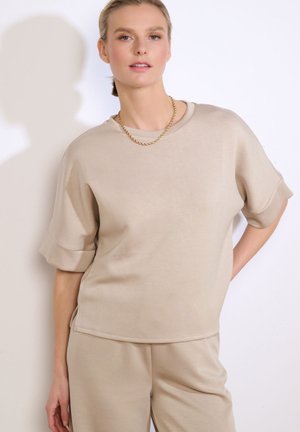 Mujer con camiseta de manga corta beige y pantalones, complementada con un collar de cadena dorada, posando frente a un fondo blanco liso.