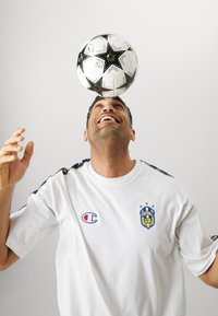 Un ballon de soccer noir et blanc équilibré sur la tête d'un homme portant un t-shirt blanc avec un logo et des accents colorés sur les manches.