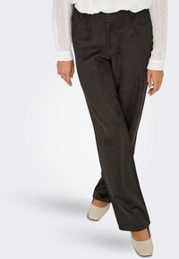 Femme portant un pantalon noir droit, un chemisier blanc à manches longues transparentes et des chaussures beiges à bout carré, debout jambes croisées.