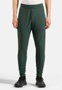 Mann in dunkelgrünen, körperbetonten Sporthosen und schwarzen Laufschuhen, der mit entspannten Armen an den Seiten vor einem weißen Hintergrund steht.