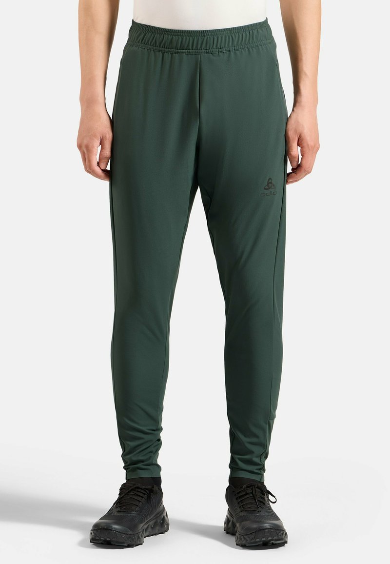 Mann in dunkelgrünen, körperbetonten Sporthosen und schwarzen Laufschuhen, der mit entspannten Armen an den Seiten vor einem weißen Hintergrund steht.