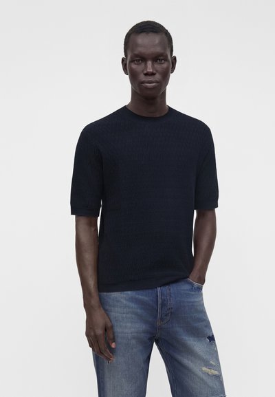 Homme debout avec une main dans la poche, portant un pull noir à manches courtes et un jean bleu déchiré sur un fond uni.