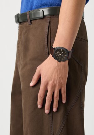 De linkerhand van een man die langs zijn zij rust, draagt een zwartbruine polshorloge, bruine broek, zwarte riem en blauwe blouse.