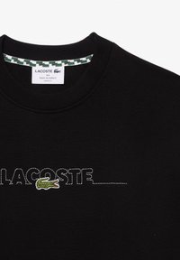 Sudadera negra con cuello redondo, corte holgado, que presenta el texto "LACOSTE" bordado en blanco y un logo de cocodrilo verde a la izquierda.