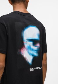 Fekete pamut póló, élénk grafikai mintával, amelyen egy torzított arc látható kék, rózsaszín és fehér színekben, a "KARL LAGERFELD JEANS" felirattal.