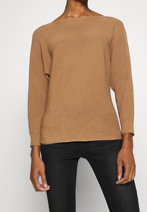 Person iført en kamelfarvet ribstrikket sweater med lange ærmer og sorte tætsiddende bukser, stående mod en ensfarvet baggrund.