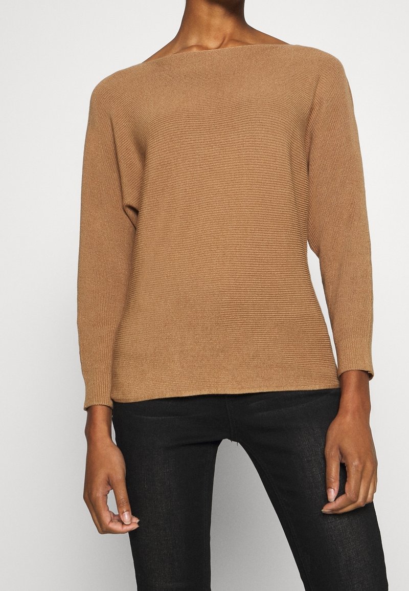 Personne portant un pull en maille côtelée couleur camel à manches longues et un pantalon noir ajusté, debout devant un fond uni.
