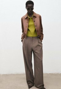 Giacca in pelle marrone sopra un maglione a coste verde, abbinata a pantaloni a gamba larga a quadri marroni. Occhiali da sole oversize completano il look.
