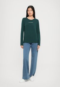 Kobieta ubrana w ciemnozielony longsleeve z białym logo, szerokie niebieskie dżinsy i białe trampki, stojąca na tle jednolitego tła.
