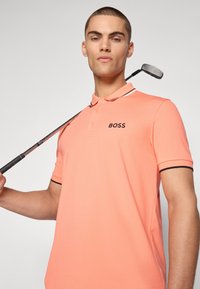 Polo shirt corallo con logo ricamato "BOSS" nero, finiture a contrasto nere su colletto e maniche; il modello tiene una mazza da golf.