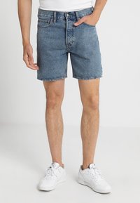 Shorts de mezclilla en azul claro, con un corte clásico, diseño de cinco bolsillos y cierre de botón. Llevados con zapatillas blancas.