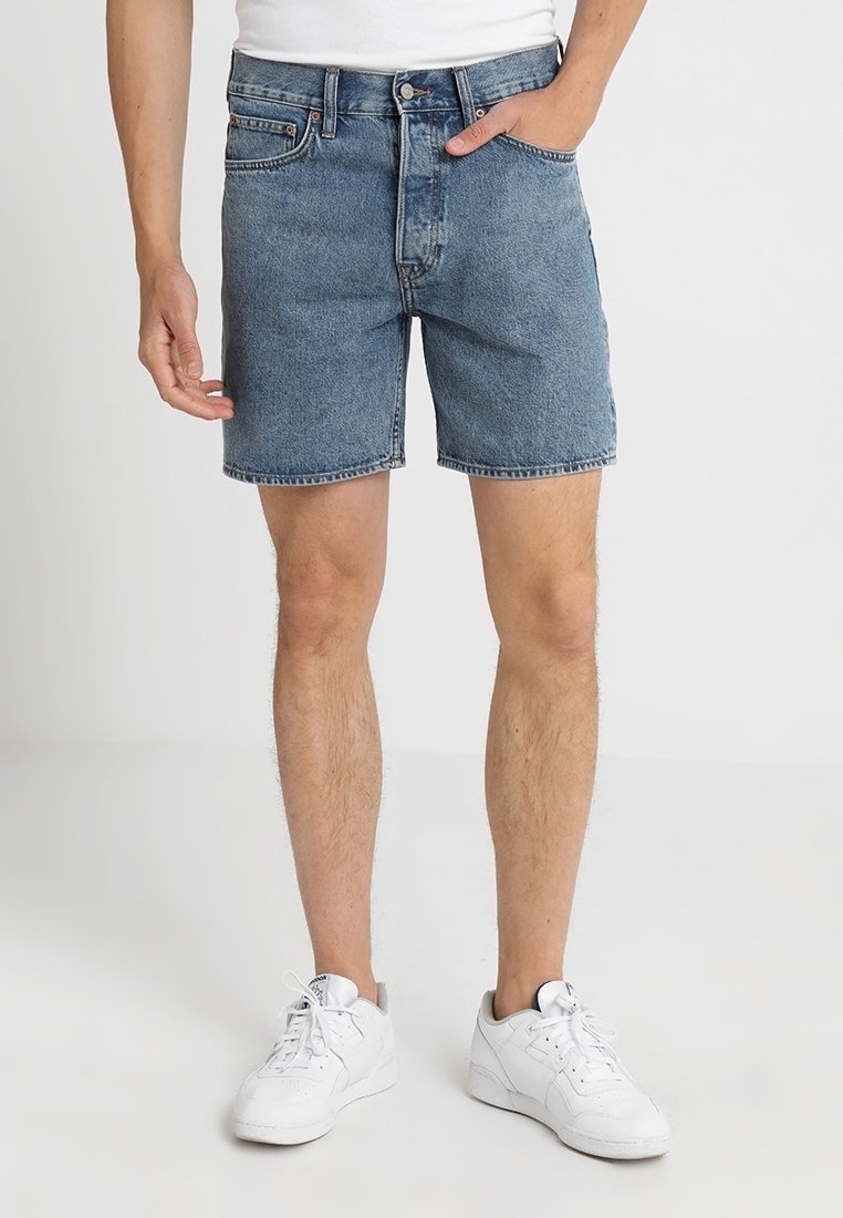 Shorts de mezclilla en azul claro, con un corte clásico, diseño de cinco bolsillos y cierre de botón. Llevados con zapatillas blancas.