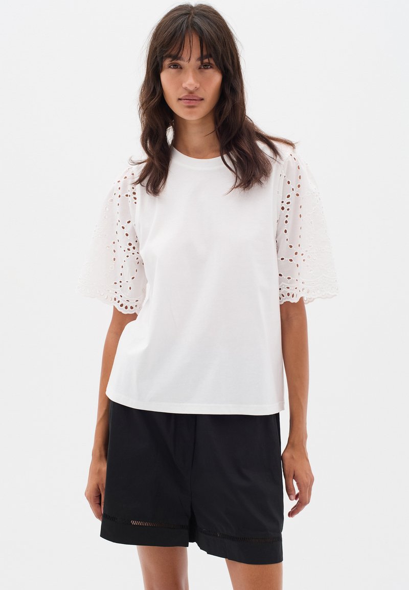 T-shirt bianco in cotone con maniche in pizzo ad occhio ricamato, scollatura rotonda e vestibilità ampia, abbinato a pantaloni corti neri con orlo decorativo.