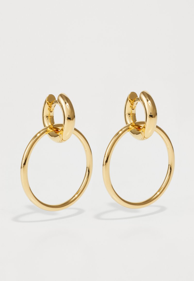 Boucles d'oreilles en métal doré, présentant une grande forme circulaire dotée d'une texture lisse et polie, avec un accent en forme d'ovale en haut.
