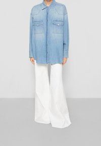 Světle modrá denimová košile s dlouhými rukávy, špičatým límcem a dvěma náprsními kapsami, spojená s bílými širokými kalhotami na neutrálním pozadí.