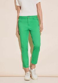 Gröna, åtsittande bomullsbyxor med medelhög midja, sidofickor och lätt stretch; bärs med vita sneakers och en texturerad vit topp.