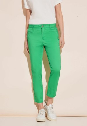 Pantaloni in cotone verdi, aderenti, con vita media, tasche laterali e una leggera elasticità; indossati con sneakers bianche e un top bianco texturizzato.