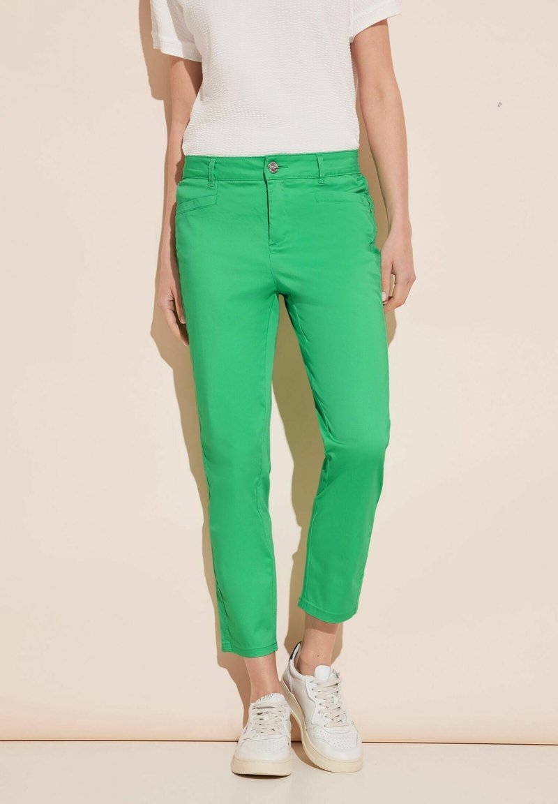 Gröna, åtsittande bomullsbyxor med medelhög midja, sidofickor och lätt stretch; bärs med vita sneakers och en texturerad vit topp.