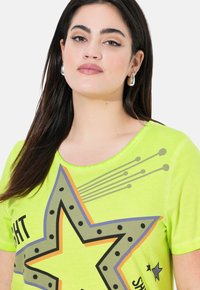 Lys grøn kortærmet t-shirt med et stort stjernedesign med sorte prikker og orange accenter, rund halsudskæring og blødt stof.