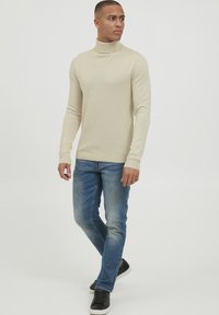 Bež turtleneck pulover iz mehkega blaga, v kombinaciji s prilagojenimi modrimi kavbojkami in črnimi supergami. Enostaven in sodoben dizajn.