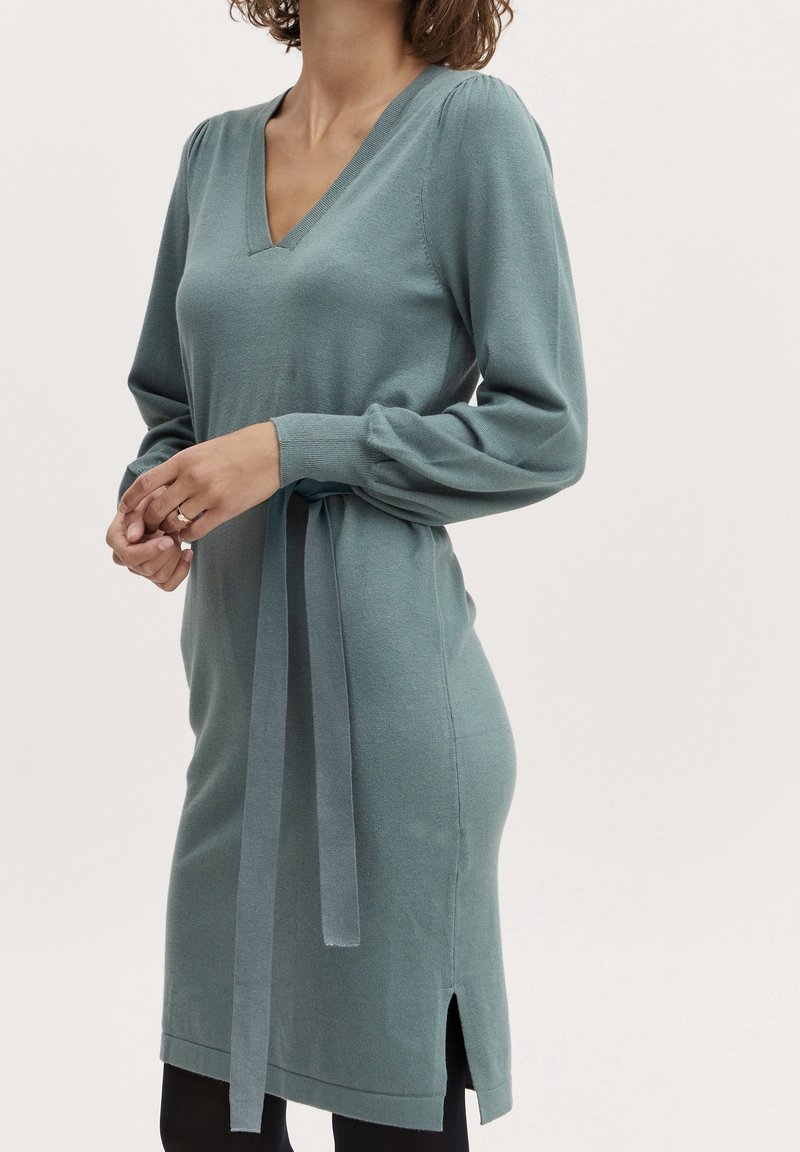 Abito lavorato a maglia color teal con scollo a V, maniche lunghe a sbuffo, vita regolabile con laccetto e spacco laterale all'orlo. Realizzato in tessuto morbido con una texture liscia.