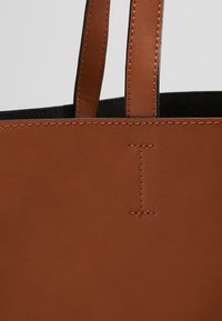 Sac fourre-tout en cuir marron avec deux poignées et des détails de couture visibles sur la surface lisse et les anses, sur un fond clair.