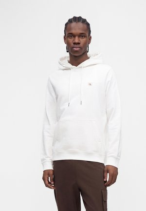 Giovane uomo con capelli intrecciati che indossa una felpa bianca con cappuccio con un piccolo logo e pantaloni marroni su uno sfondo chiaro e uniforme.