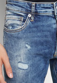 Jeans de mezclilla azul con un diseño desgastado, que incluye roturas, parches más claros, costuras de cobre y un estilo clásico de cinco bolsillos.