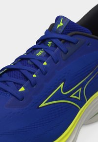 Μπλε αθλητικό παπούτσι με λογότυπο και λεπτομέρειες σε νέον κίτρινο, με λεπτομερή επάνω μέρος από δίχτυ και σφιχτά δεμένα κορδόνια σε λευκό φόντο.