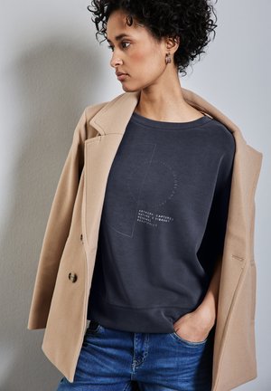 Dunkelgrauer Sweatshirt mit rundem Ausschnitt, versehen mit einem weißen Textdesign. Kombiniert mit einem beigen Mantel, der eine strukturierte Silhouette präsentiert.