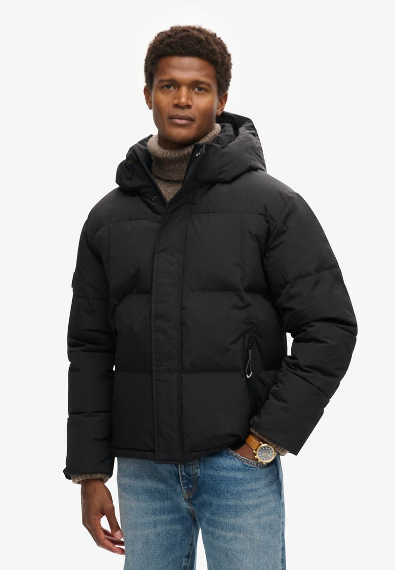 Giacca a vento nera con cappuccio, con sezioni trapuntate, tasche laterali e chiusura con zip, indossata su un dolcevita color marrone chiaro.