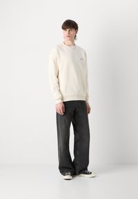 Sudadera color crema con puños y bajo de canalé, que presenta un pequeño logo; combinada con unos vaqueros de pierna ancha gris oscuro y zapatillas negras.