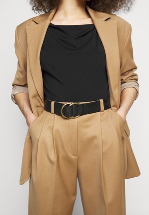 Femme portant un costume couleur camel avec manches retroussées, top noir et large ceinture noire avec boucle double anneau dorée, mains dans les poches.