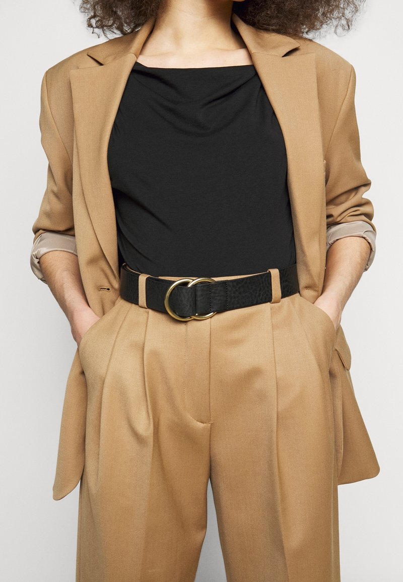 Mujer con traje color camello con mangas remangadas, top negro y cinturón ancho negro con hebilla dorada de doble anillo, manos en los bolsillos.