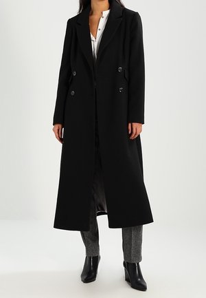 Femme portant un long manteau noir croisé par-dessus une chemise blanche, un pantalon gris et des bottines noires, debout devant un fond uni.
