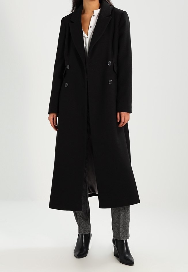 Femme portant un long manteau noir croisé par-dessus une chemise blanche, un pantalon gris et des bottines noires, debout devant un fond uni.