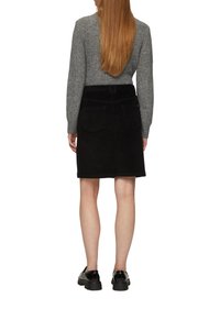 Schwarzer Cordrock, knielang, mit aufgesetzten Taschen hinten und leicht ausgestelltem Design. Getragen mit einem grauen Strickpullover und schwarzen Schuhen.